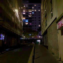 Narrow street ends with a tall apartment building with multiple colors of lights coming out of the windows.
