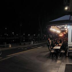 People sit by a well lit crowded bar. The background is completely black in comparison. A cyclists is going by, showing the red light on the back.