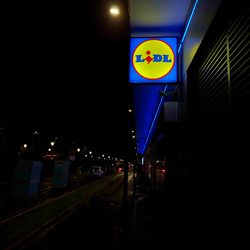 Blue and yellow Lidl sign shining in the foreground on a dark boulevard.