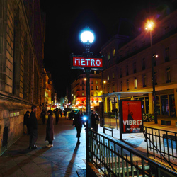 A red Metro entrance sign post. Behind there are people heading towards the stairs and further behind are the lights of a busy commercial street. The metro lightpost gives off a white blueish light white the street lights are orange.