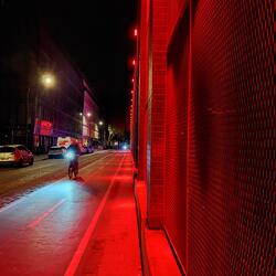 Bike lane along the side of a building that has red lights, making the entire lane bathed in red. One cyclist is riding away from the camera in the foreground. In the background, just by the light you can see another bike coming in the direction of the camera.