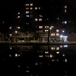 Lit up mid-rise building with it's window lights reflected in the still water it sits next to.