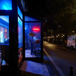 Blue light from the windows of a building. There is a red neon sign that says "SANDWICH". Two people on the side are seen walking down the street next to it.