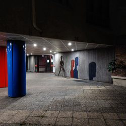The grey entrance of a building. There is a statue of a humanoid shape in place. Behind is what look like red, blue, and black shadows of human heads. There is a blue column and red column holding up the structure.