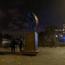Statue with blue colored woman athlete. Engraving: Les 4 Cardinales du Sports. Kuntu, "ouest" en quechua. Olympiade Culturelle 2024. Ville de Paris. Silhouettes of two men are chatting by the statue. Behind are lit up buildings along the Canal Saint Martin.